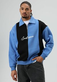 Sweat-shirt à fermeture éclair bleu et noir avec un col haut, présentant des panneaux contrastants et le texte "Sautessence" en cursive blanche.