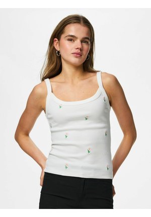 Jeune femme aux longs cheveux, portant un débardeur blanc avec de petites fleurs brodées et un pantalon noir, les mains posées sur les hanches.
