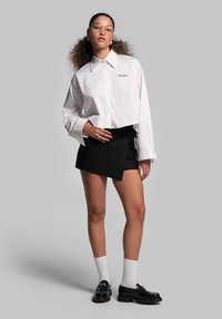 Weißes, kurzes Hemd mit langen Ärmeln und Kragen, mit schwarzer Stickerei verziert. Kombiniert mit einem schwarzen asymmetrischen Minirock und Loafers.