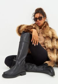 Black leather thigh-high boots with a sleek design, round toe, and low block heel, worn with a faux fur coat and black leggings.