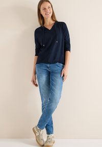 Haut à capuche manches longues bleu marine associé à un jean bleu clair et des baskets beige. Le haut présente un col en V et des accents à cordon de serrage.