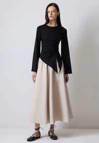 Maglietta nera a maniche lunghe con arricciature e dettaglio a fiocco, abbinata a una gonna beige fluida e sandali neri con lacci, il tutto su uno sfondo chiaro.