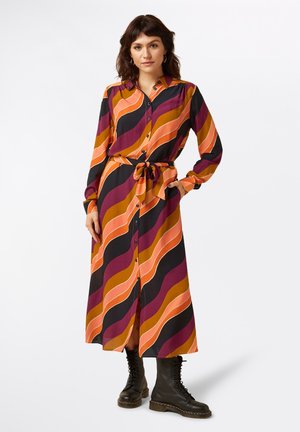 Knielanges Hemdblusenkleid mit gewellten Streifen in Orange, Lila und Schwarz, tailliert mit einem Gürtel, langen Ärmeln und knöchelhohen schwarzen Stiefeln.