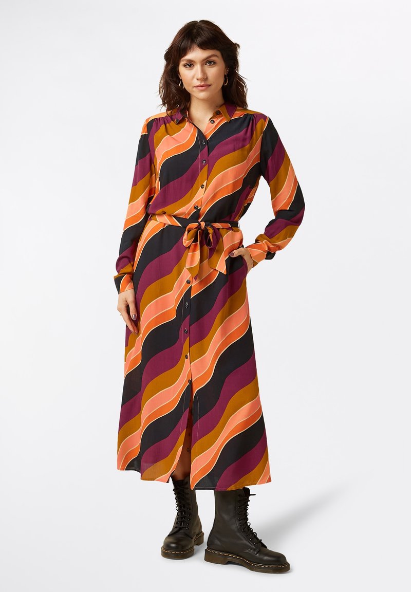 Knielanges Hemdblusenkleid mit gewellten Streifen in Orange, Lila und Schwarz, tailliert mit einem Gürtel, langen Ärmeln und knöchelhohen schwarzen Stiefeln.
