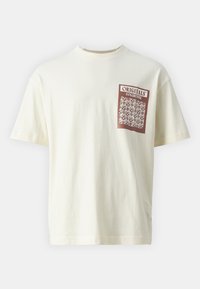 T-shirt couleur crème en coton avec un imprimé graphique marron sur la zone gauche de la poitrine, incluant du texte et des motifs complexes. Manches courtes.
