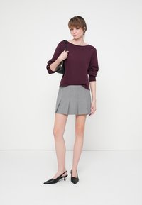 Kobieta stoi, ubrana w burgundowy sweter, szarą plisowaną spódnicę, czarne buty na obcasie typu slingback i trzyma czarną torebkę na ramieniu.