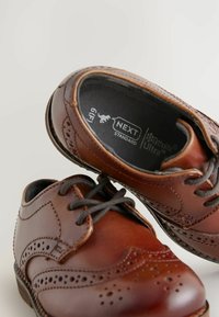 Brązowe skórzane buty brogue mają szczegółowe perforacje, okrągły czubek, teksturowane sznurowadła oraz gładką powierzchnię. Wewnątrz znajduje się etykieta "NEXT Standard."