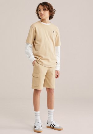 Dreng med skulderlangt hår står med hænderne i lommerne iført beige shorts og t-shirt over en hvid langærmet skjorte samt hvide sneakers.