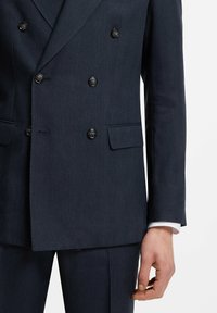 Mann trägt eine dunkelmarineblaue zweireihige Anzugjacke mit schwarzen Knöpfen und passende Hose vor einem einfarbigen Hintergrund.