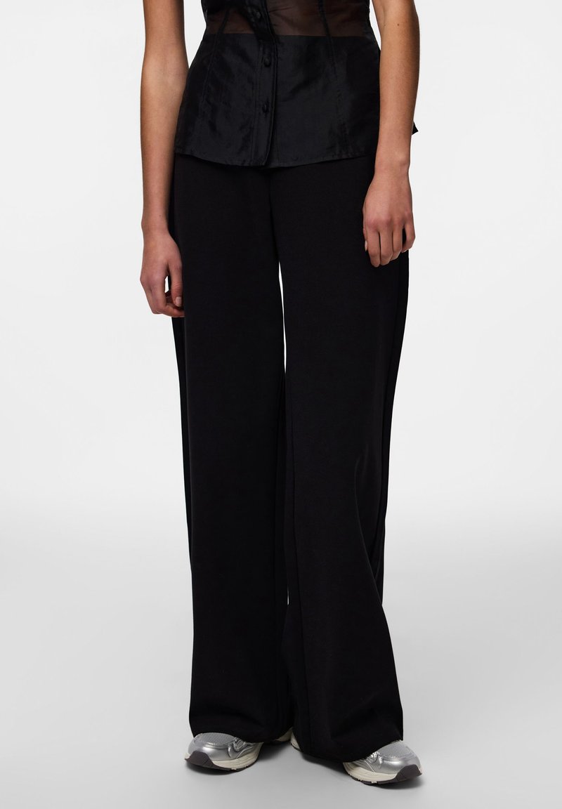 Črne široke hlače iz mehkega blaga, kombinirane s črnim prosojnim oprijetim topom s detajli gumbov. Model nosi srebrne superge.