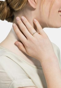 Goldring mit einem kleinen zentralen Edelstein, getragen an einer Hand, die sanft den Hals berührt. Glatte Textur, zartes Design und schlanke Bandform.