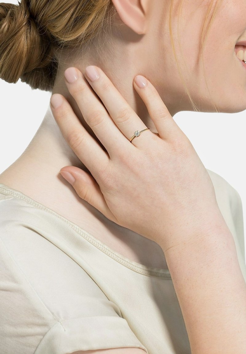 Goldring mit einem kleinen zentralen Edelstein, getragen an einer Hand, die sanft den Hals berührt. Glatte Textur, zartes Design und schlanke Bandform.