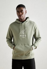 Haut à capuche vert olive en coton, présentant une poche avant et un logo avec des ailes. Coupe décontractée avec cordon de serrage ajustable au niveau de la capuche.