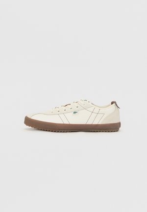 Weißer Sneaker in niedrigem Schnitt mit brauner Gummisohle und Schnürsenkeln, seitliche Nähte und ein kleines grünes Logo-Tag.