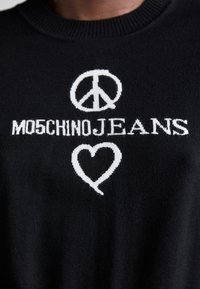 Melnā adīta džemperis ar baltiem uzrakstiem "MOSCHINO JEANS", miers simbolu un sirds dizainu, uzsverot kontrastu un faktūru.