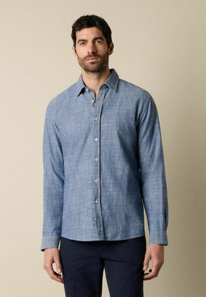 Man met donker haar draagt een lichtblauw overhemd met lange mouwen en donkere broek, staande tegen een effen beige achtergrond.