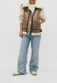 Hnědá umělá kůže vesta s krémovým ovčím límcem, se zipem vpředu, bočními kapsami a hnědými akcenty. Spárováno se světle modrými džíny a teniskami.