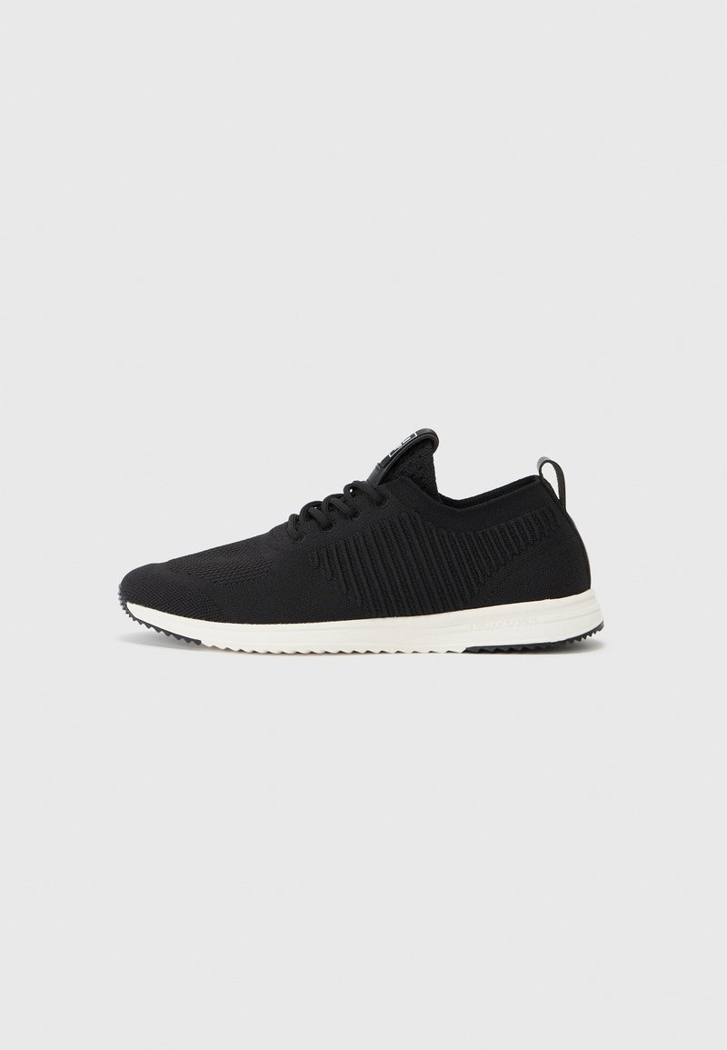 Pantof de sport negru cu un superior din tricot texturat, talpă albă și un mâner la călcâi. Prezintă elemente de design minimalist.
