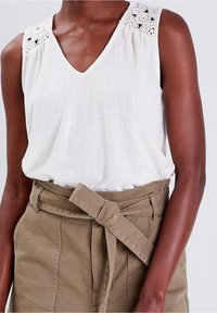 Haut sans manches blanc avec un col en V et des détails en dentelle complexes sur les épaules, associé à un pantalon ceinturé vert olive.