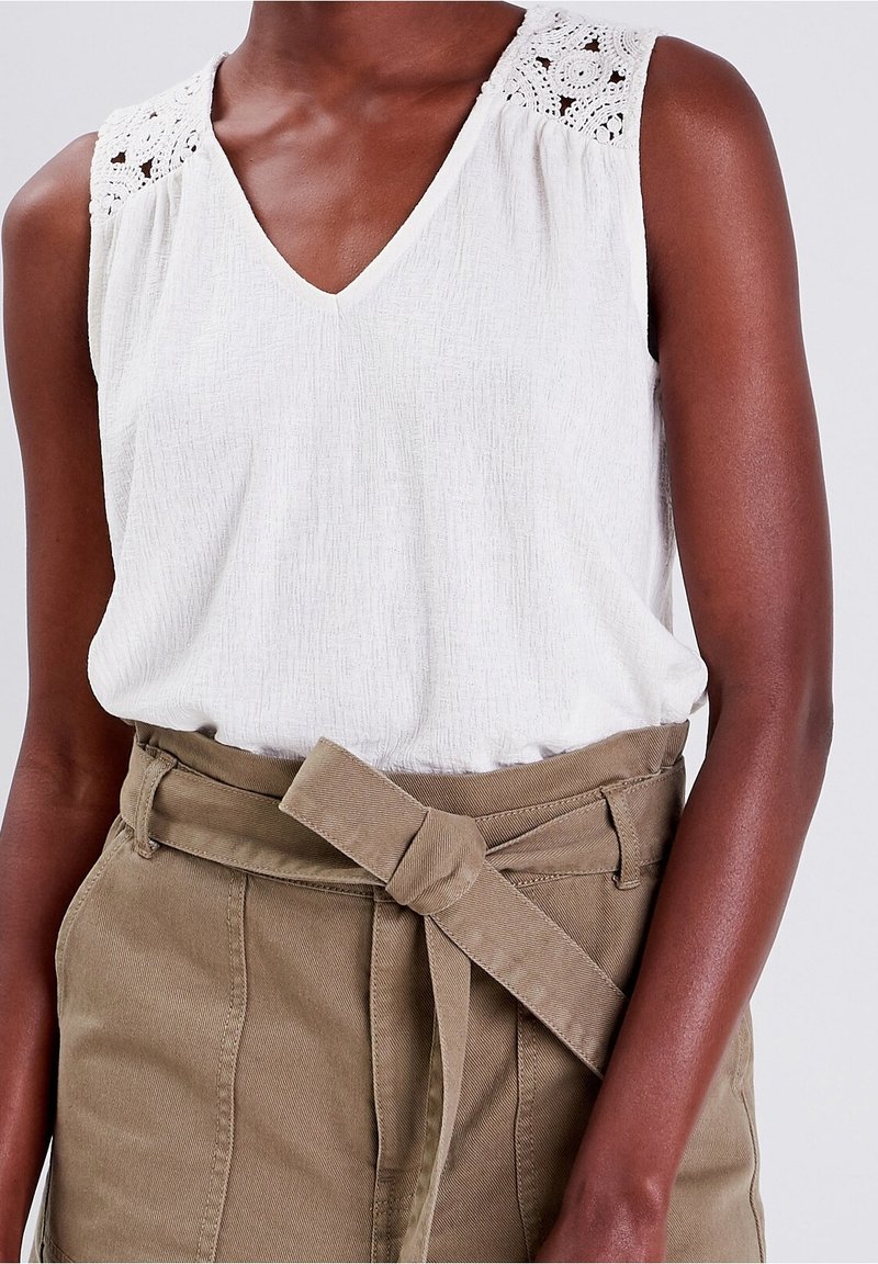 Haut sans manches blanc avec un col en V et des détails en dentelle complexes sur les épaules, associé à un pantalon ceinturé vert olive.