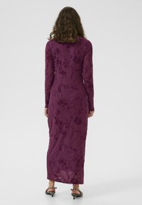 Kvinna med vågigt hår som bär en långärmad, golvlång lila klänning med blommotiv, stående vänd bort från kameran mot en enfärgad bakgrund.