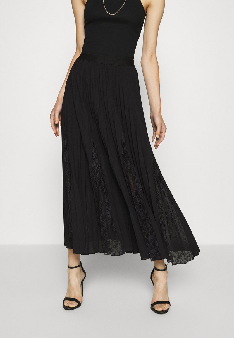 Femme portant une jupe longue noire plissée avec des inserts en dentelle, un top noir sans manches, des sandales à talons hauts avec bride à la cheville et un collier fin en or.