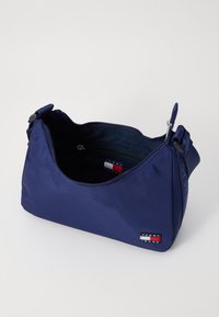 Sac à épaule en nylon bleu marine avec une forme incurvée, fermeture éclair et bandoulière réglable. Présente un patch logo Tommy Hilfiger sur le devant.