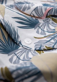 Chemise à manches courtes avec un fond blanc et un motif de feuilles tropicales bleues, vertes et roses. Présente un bouton blanc et une texture lisse.