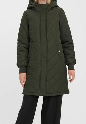 Femme portant un long manteau d'hiver matelassé vert foncé avec une capuche, des poches latérales et un pantalon noir, sur un fond uni.