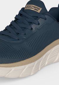 Σκούρο μπλε αθλητικό παπούτσι Skechers BOBS με υφασμάτινο επάνω μέρος με υφή, κορδόνια ίδιου χρώματος, μπεζ ενδιάμεση σόλα και λευκή σχέδια στην εξωτερική σόλα.