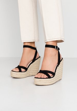 Czarne sandały na koturnie z zamszu z plecioną platformą z juty i paskiem wokół kostki; liczne krzyżujące się paski na palcach, gładka wewnętrzna wyściółka.