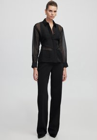 Camicia nera trasparente a maniche lunghe con una trama arricciata, abbottonata sul davanti e dal taglio strutturato, abbinata a pantaloni neri a vita alta.