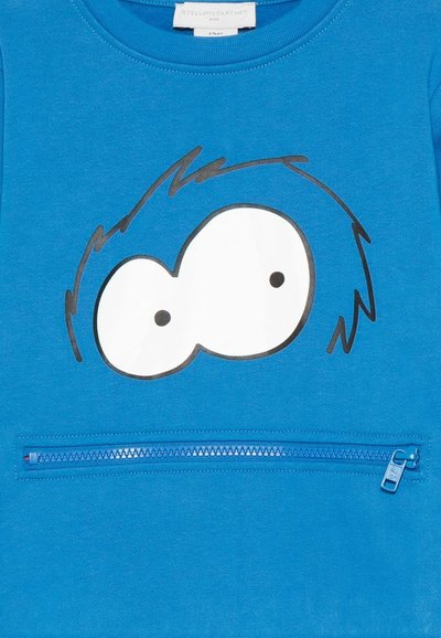 Maglietta blu per bambini con un disegno di occhi da cartone animato e capelli neri sopra una tasca verticale blu con cerniera.