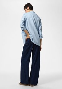 Persoon draagt een lichtblauw shirt met lange mouwen en wijd uitlopende donkerblauwe spijkerbroek, staat met één hand in de achterzak.