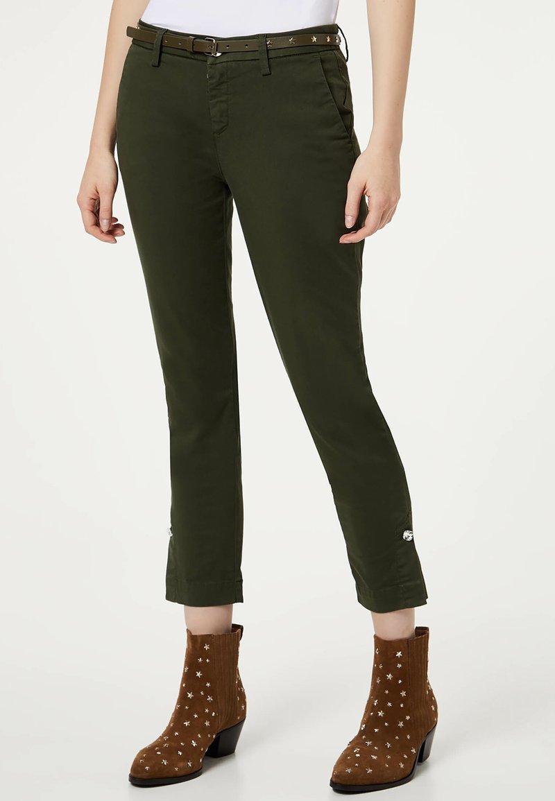 Femme portant un pantalon court vert foncé avec des détails de boutons aux poignets, une fine ceinture marron et des bottines en daim marron avec des clous en forme d'étoile.