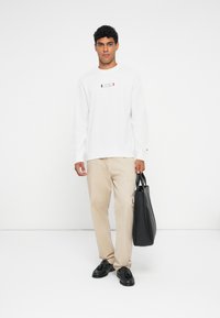 Valge sviiter logoga; beežid püksid; hoiab musta nahast kottidekotti. Sile kangas, lained sobivus, minimalistlik disain. Mustad kingad.