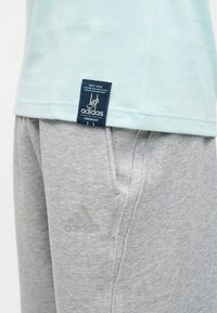 Svetlo modra majica Adidas s črnim oznako blizu roba in svetlo sive Adidas trenirke z bočnim žepom in logotipom.