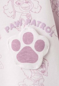 Vêtement violet clair avec un patch en forme d'empreinte de patte blanche aux accents roses et le texte "PAW PATROL", sur un fond à motif répétitif subtil de personnages.
