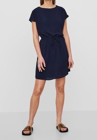 Tmavě modré šaty s krátkými rukávy a volným střihem, stažené v pase pomocí šňůrky. Nošeny s plochými sandály s otevřenou špičkou.