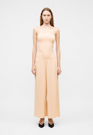 Femme aux longs cheveux ondulés portant un haut beige sans bretelles et un pantalon beige à jambes larges, debout devant un fond clair uni.