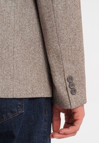 Braune Fischgratjacke mit glattem Textur an den Ärmeln. Drei dunkle Knöpfe am Bündchen. Getragen über dunklen Jeans mit kontrastierenden Nähten.