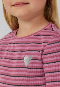 Rozā svītrainā krekla dizains ar melnām horizontālām līnijām, kuram ir sudraba sirds uzplecs. Materiāls ir riboti, ar redzamu apaļu apkakli.