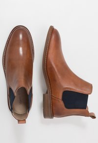 Bottes Chelsea en cuir marron avec panneaux latéraux élastiques, bout arrondi et talon bas empilé. Détails de couture le long de la semelle.