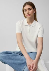 Witte polo shirt van een zachte, gladde stof met een klassieke kraag en een knoopsluiting met vijf knopen. Gecombineerd met een lichtblauwe jeansrok en witte sneakers.