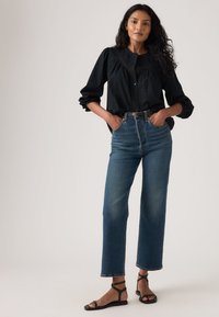 Μαύρο πουκάμισο με κουμπιά και μανίκια με όγκο, σε συνδυασμό με ψηλόμεσα μπλε τζιν και μαύρα σανδάλια με λουράκια. Ουδέτερο φόντο.