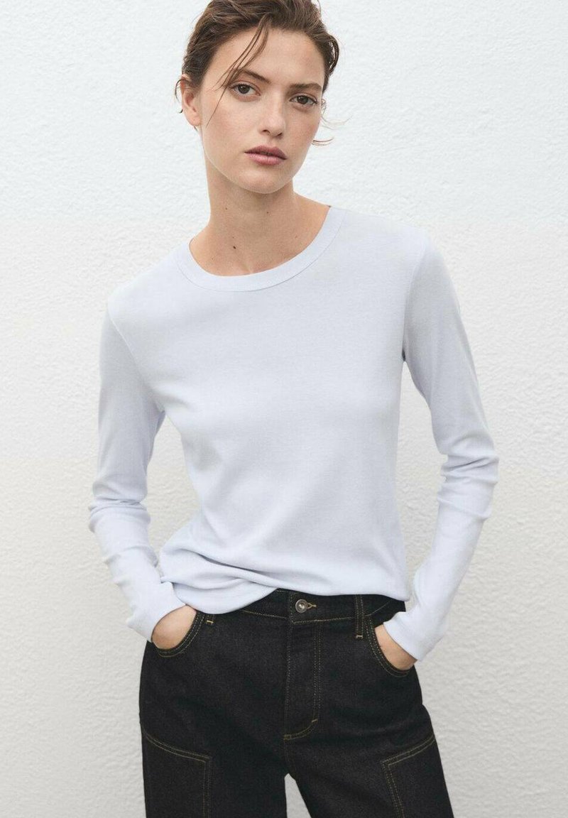 Himmelblaues Langarmhemd aus weichem Stoff, kombiniert mit dunklen Jeans mit gelben Nähten und einer eleganten Passform, Hände in den Taschen.