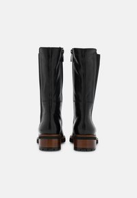 Paire de bottes montantes en cuir noir avec talons empilés marron, vues de dos sur un fond blanc uni.