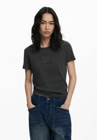 Camiseta gris carbón de manga corta, con un logo en relieve "DSGL"; combinada con jeans de mezclilla azules con grandes bolsillos delanteros.