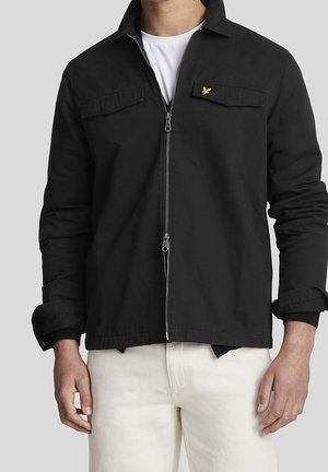 Man wearing a black zip-up jacket with two chest pockets and a small yellow bird logo on the left pocket flap, paired with white pants and a white shirt.