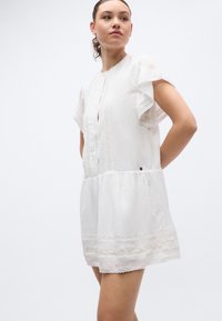 Λευκό βαμβακερό κοντό φόρεμα με ντεκολτέ σε σχήμα κλειδαρότρυπας, δαντελένιο τελείωμα, φαρδιές μανίκες, με ζωνάκι στη μέση και διακριτικές διακοσμητικές λεπτομέρειες.